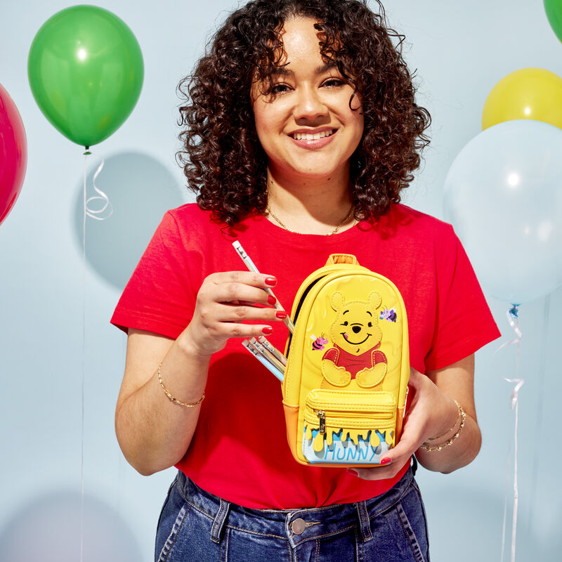 Woman with dark curly hair and wearing a red shirt holding the Loungefly Winnie the Pooh Hunny Pot Stationery Mini Backpack Pencil Case and putting pencils into it against a pale blue background with balloons 